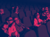 Mehrere Menschen sitzen in einem Kino, essen Popcorn und tragen 3D Brillen. Das Bild ist in magenta und lila eingefärbt. 